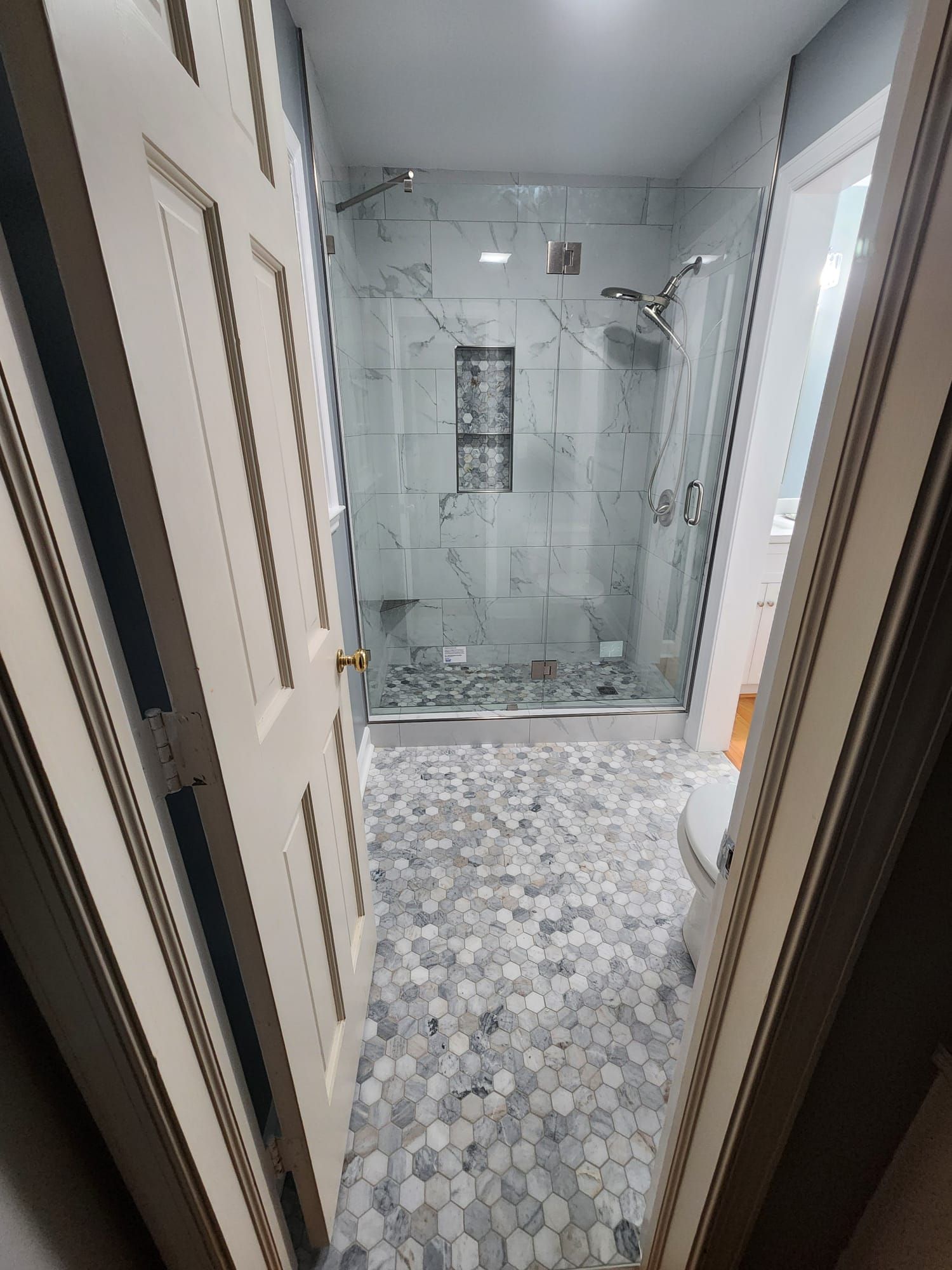 Narrow bathroom with glass-enclosed shower, mosaic tile floor, and white walls, viewed through an open doorway