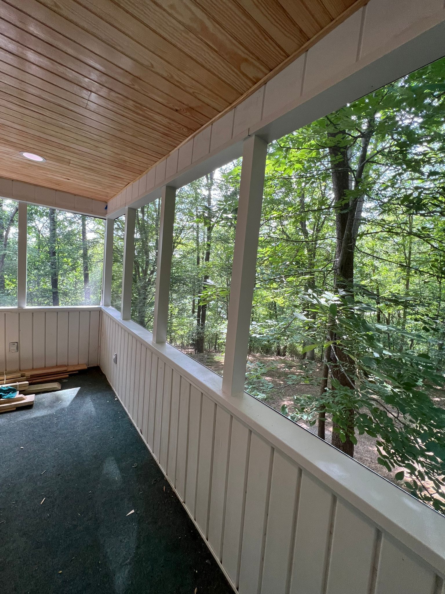 Covered wooden porch with screened railing overlooking trees, green carpeted floor, and slanted wood ceiling.