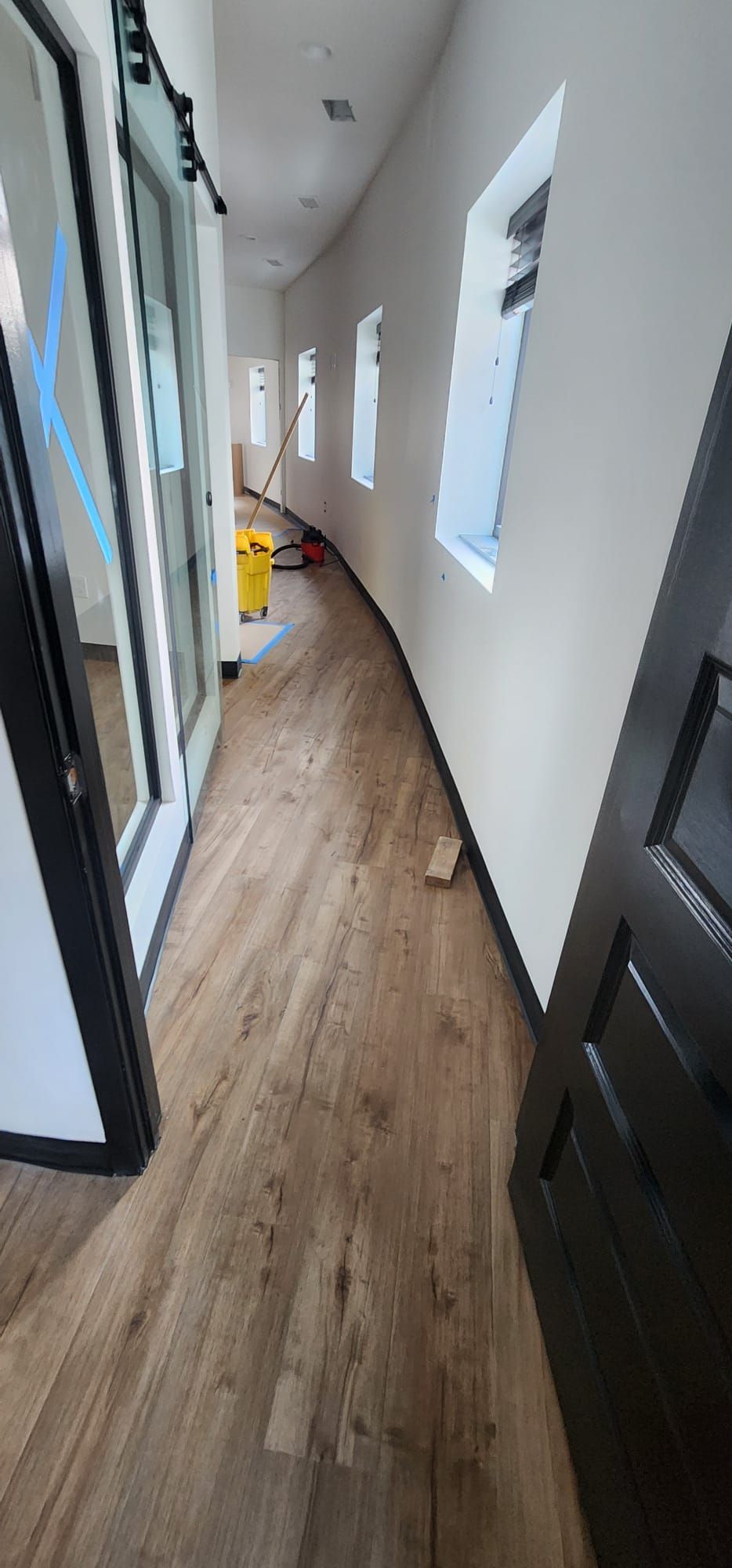 Narrow hallway with wood floors, white walls, windows, and a yellow cleaning cart in the distance