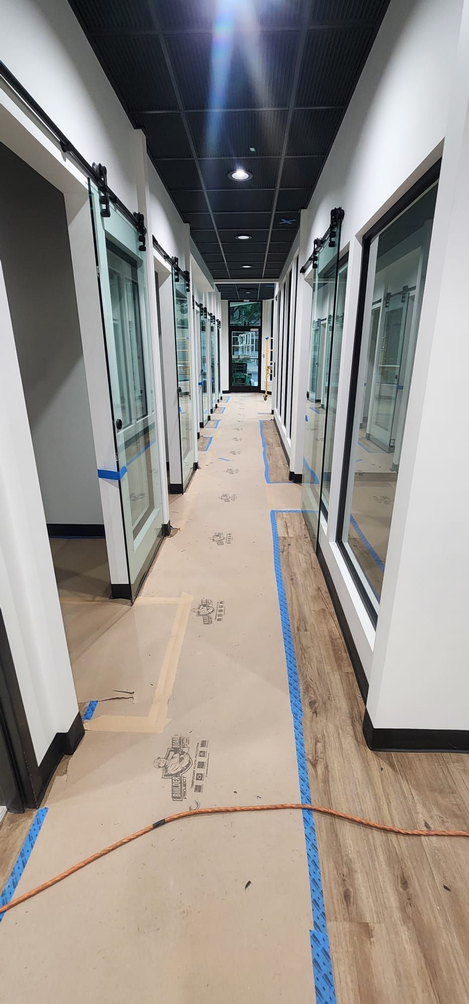 Narrow office hallway with glass-walled rooms, white walls, and a taped floor path under construction