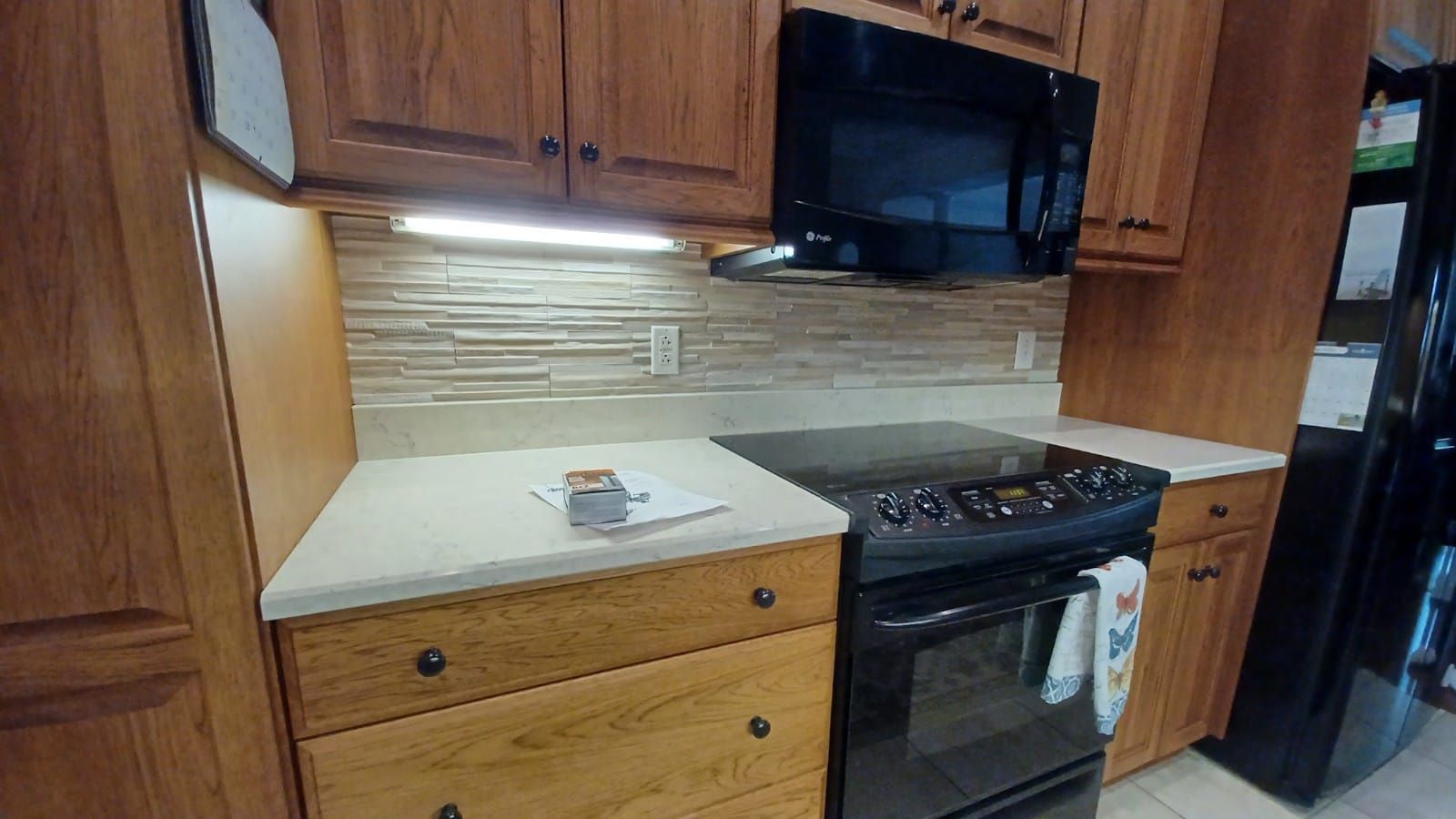 Compact kitchen with wood cabinets, black stove and microwave, beige countertops, and a black refrigerator.
