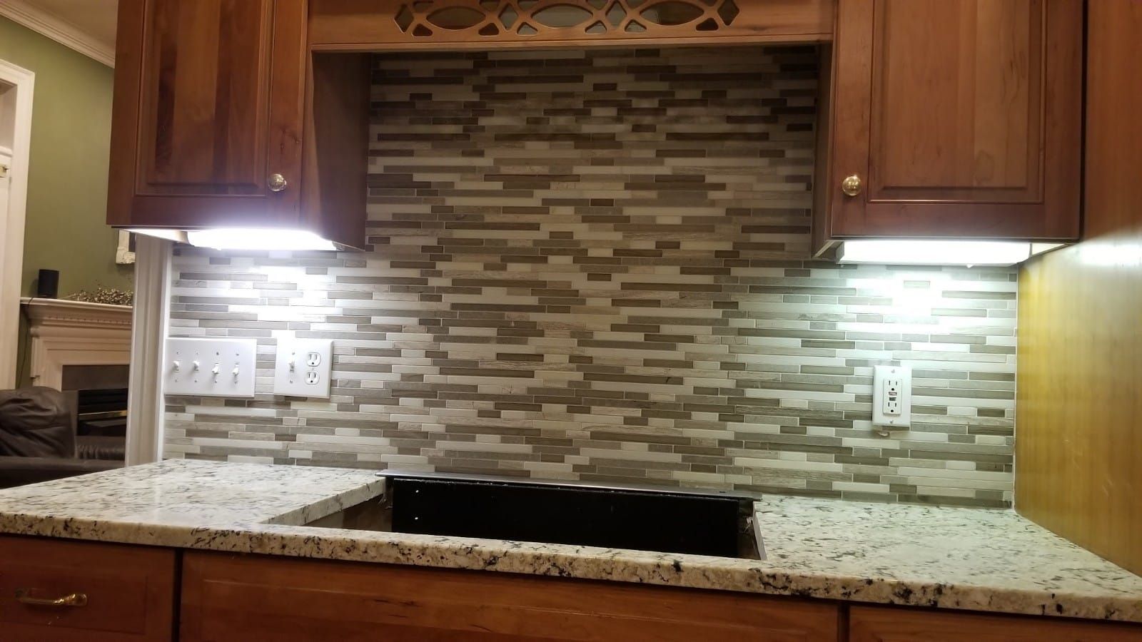 Kitchen backsplash with mosaic tile, granite countertop, and under-cabinet lights illuminating wooden cabinets.