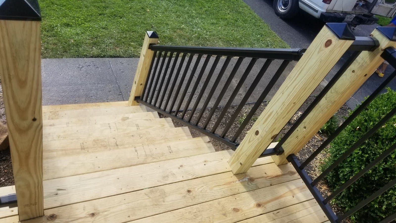 Wooden porch steps with black railings leading down to a sidewalk and grassy yard