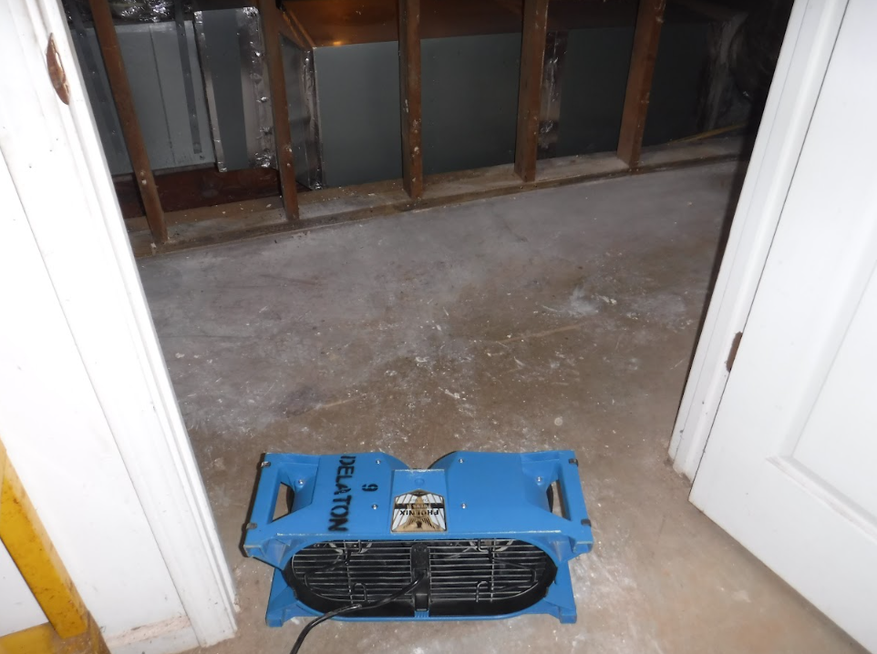 Blue fan in doorway blowing air into a damp, concrete-floored room with exposed framing and ductwork.