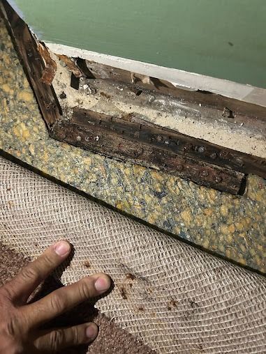 Damaged wooden frame at floor and wall junction, exposed foundation. Beige and brown tones.