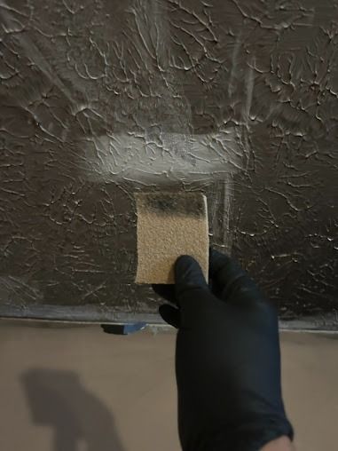 Hand in black glove holding sandpaper against dark textured surface, testing for paint removal.