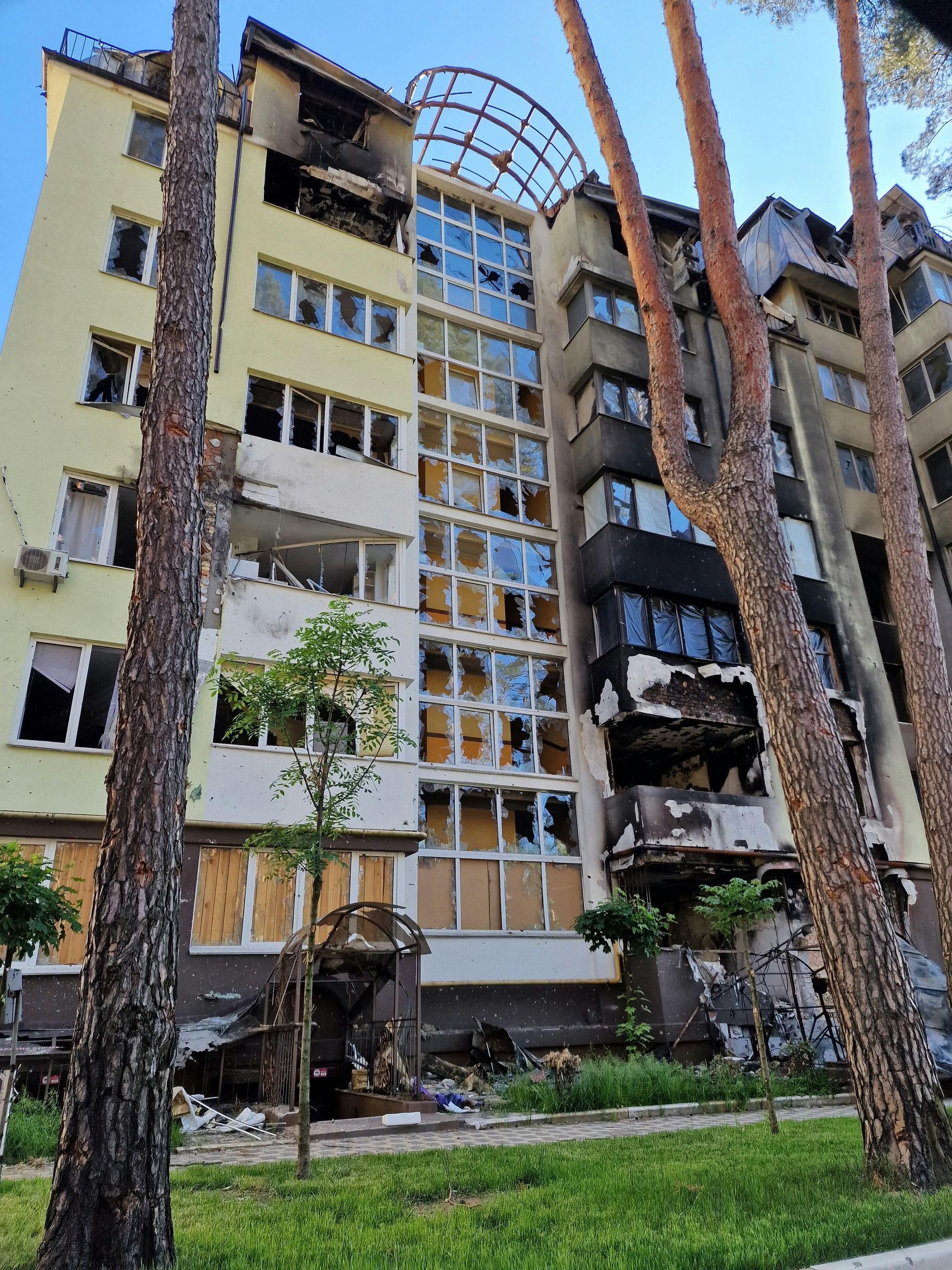 Damaged multi-story building, windows broken, charred sections, debris, green grass, and trees.