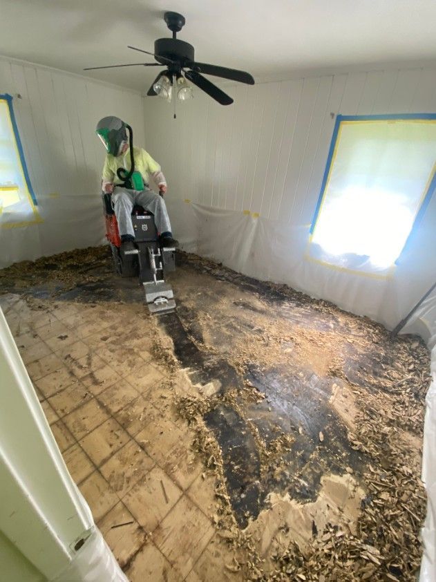 Person using a floor scraper to remove flooring in a room. Walls are covered with plastic sheeting.