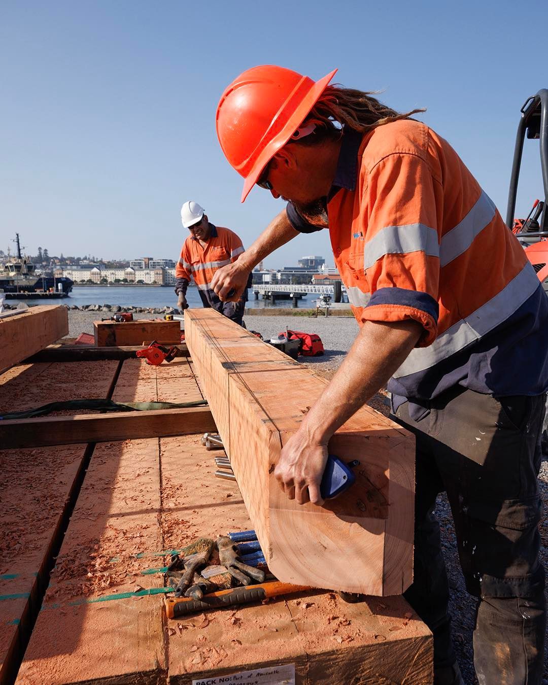 Two Workers in Orange Uniforms and Hard Hats Work with Large Timber Beams — M&J Marine Services in Medowie, NSW