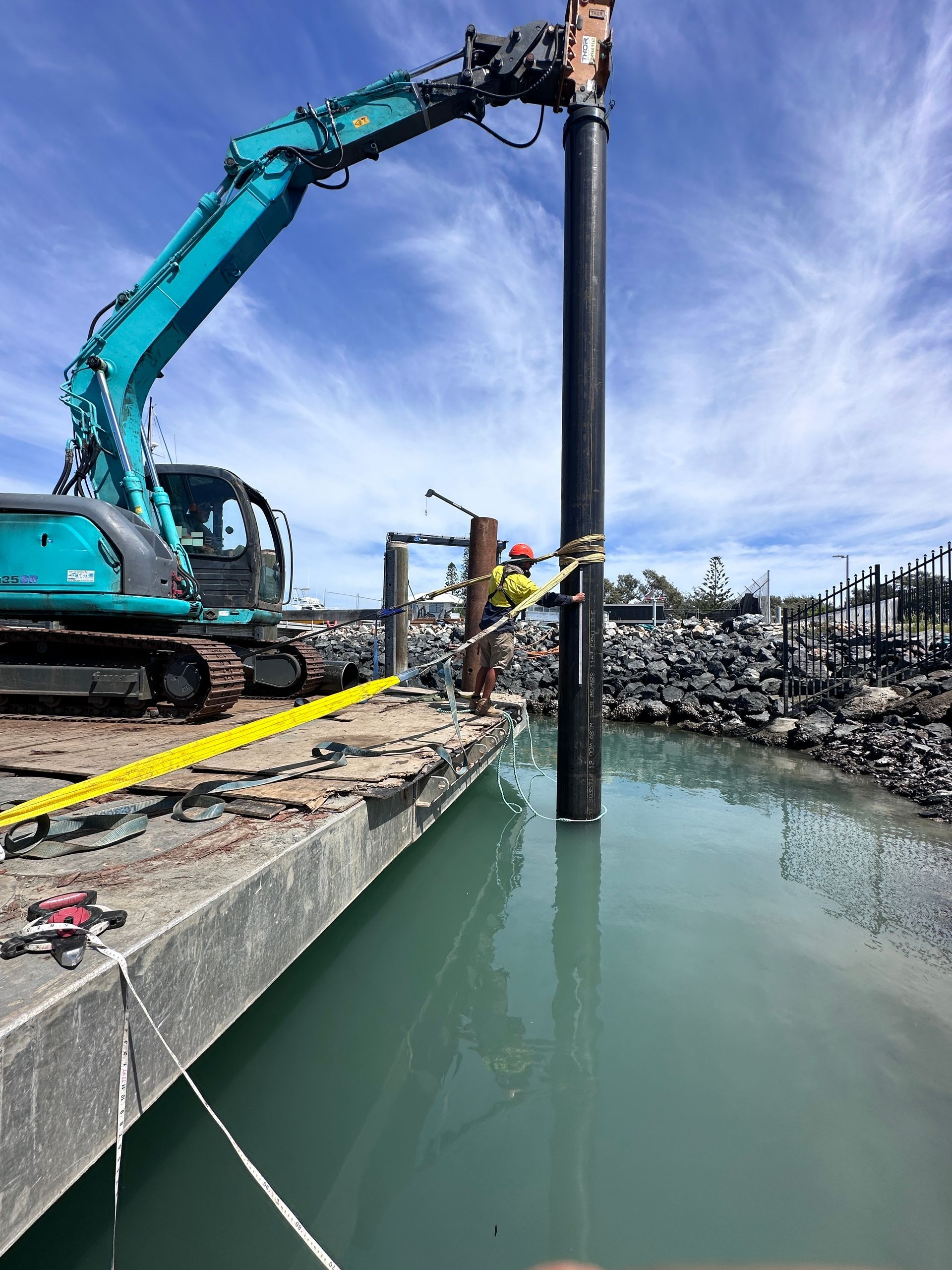 A Teal Excavator Uses an Attachment to Drive a Large Metal Piling Into the Water — M&J Marine Services in Newcastle, NSW