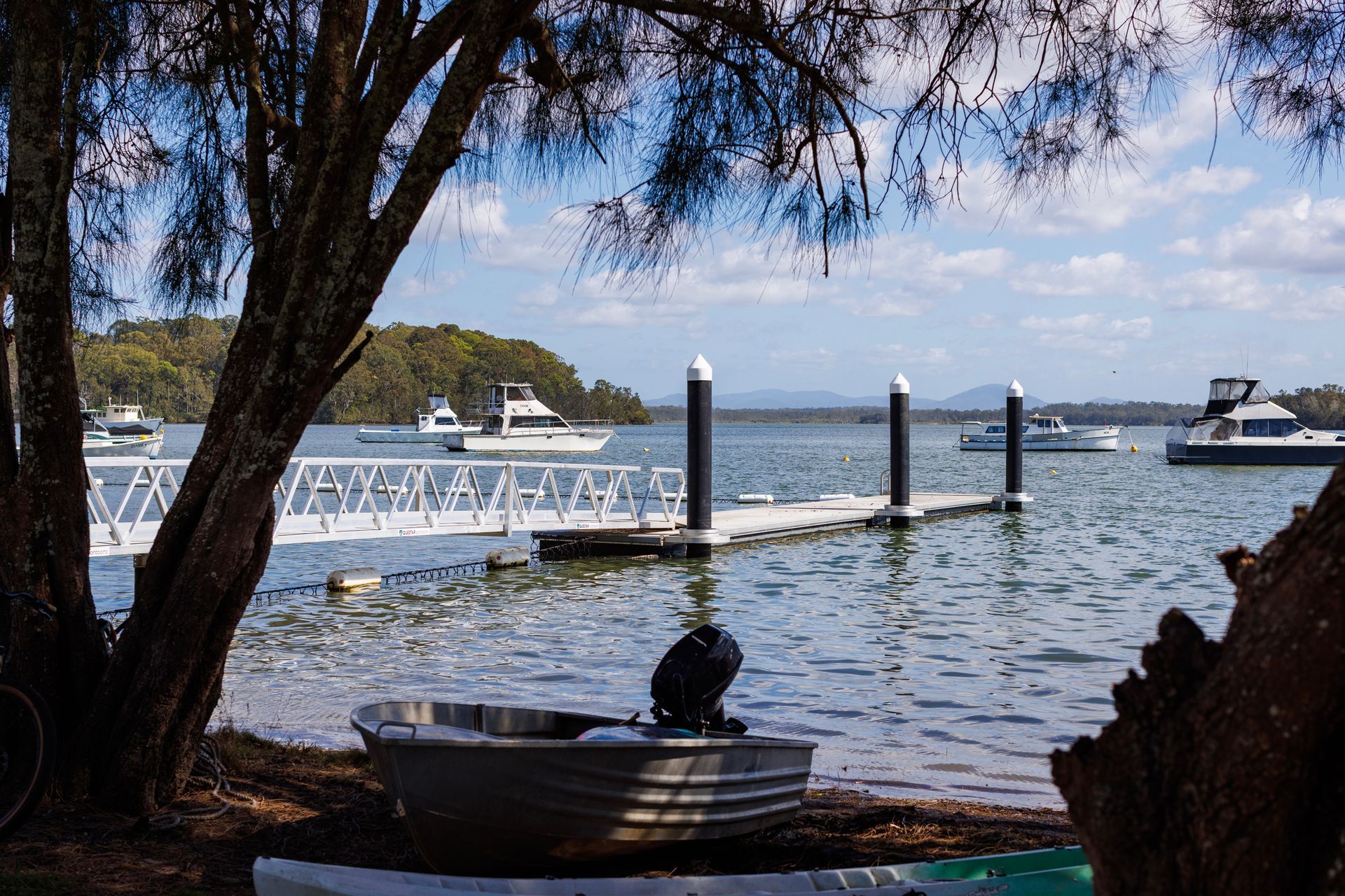 A Metal Rowboat Sits on The Shore by A Tree, with A Pier and Several Boats — M&J Marine Services in Forster, NSW
