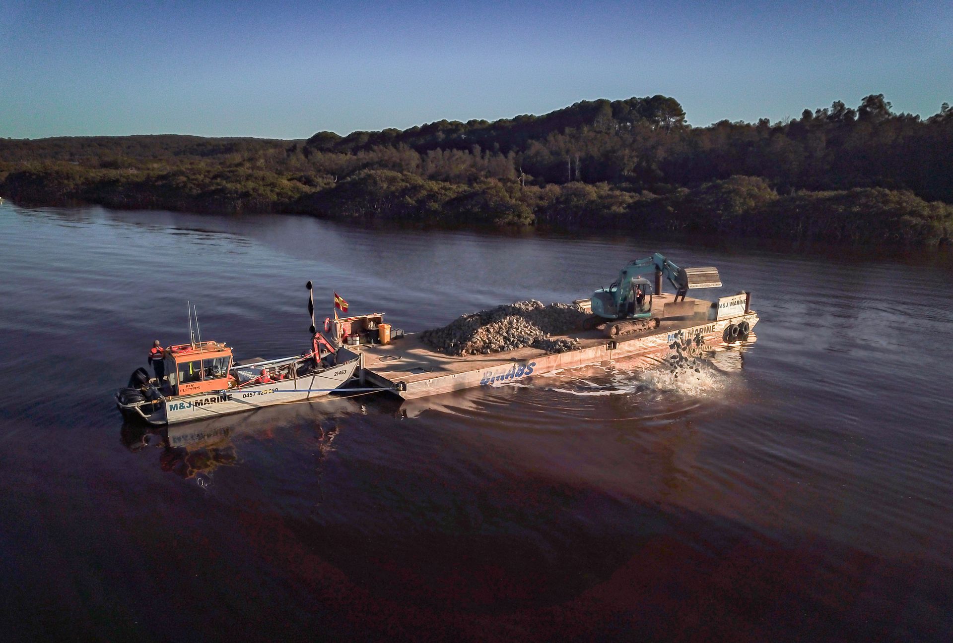 A work boat pushes a flat-deck barge carrying an excavator and piles of rocks along a calm river at sunset.