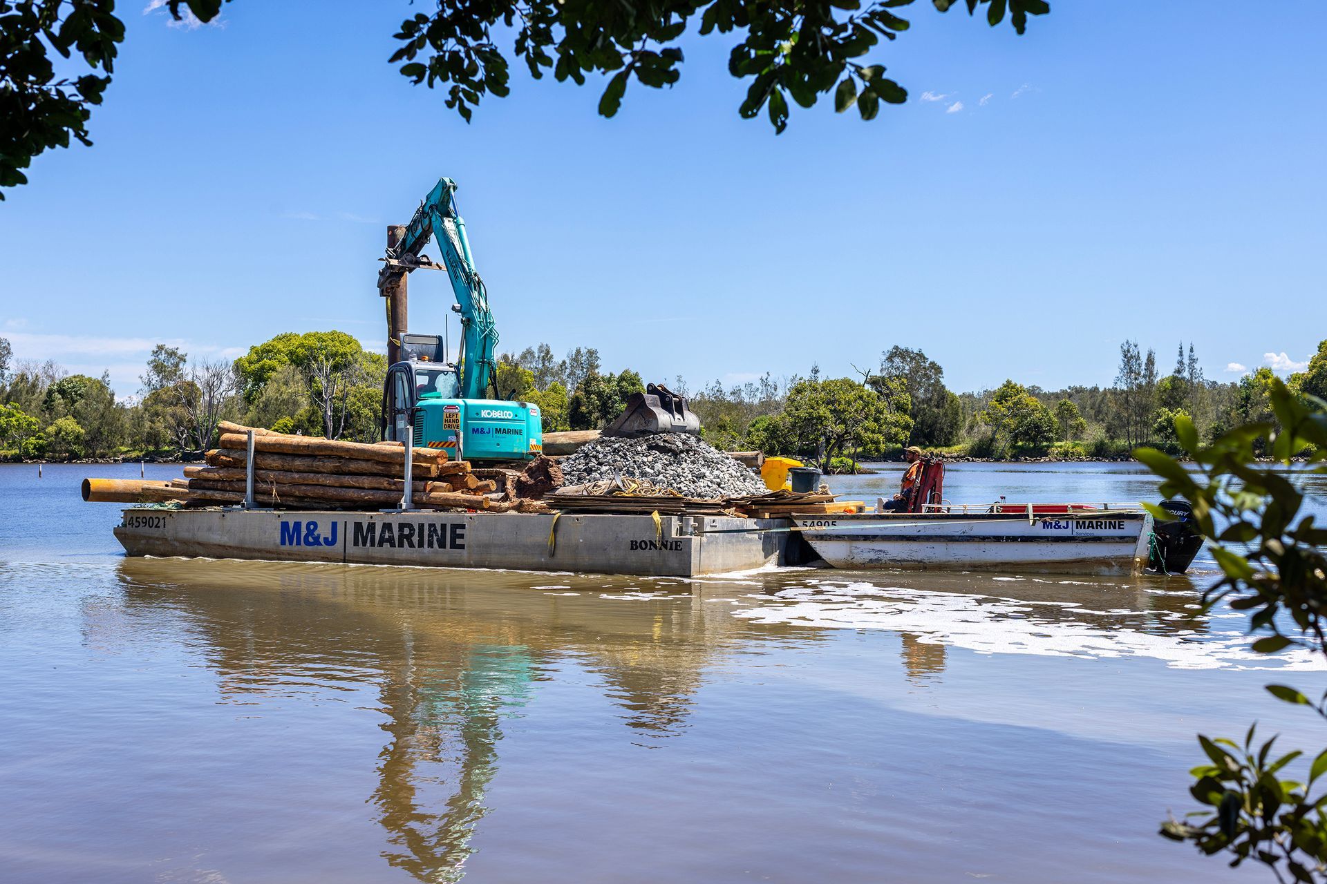 An Excavator on A Barge Carries Logs and Stones Across a Calm River — M&J Marine Services in Medowie, NSW