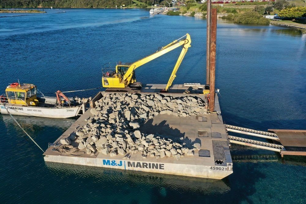 A Yellow Excavator on A Barge Is Moving Rocks in A Body of Water, Near A Wooden Dock — M&J Marine Services in Medowie, NSW