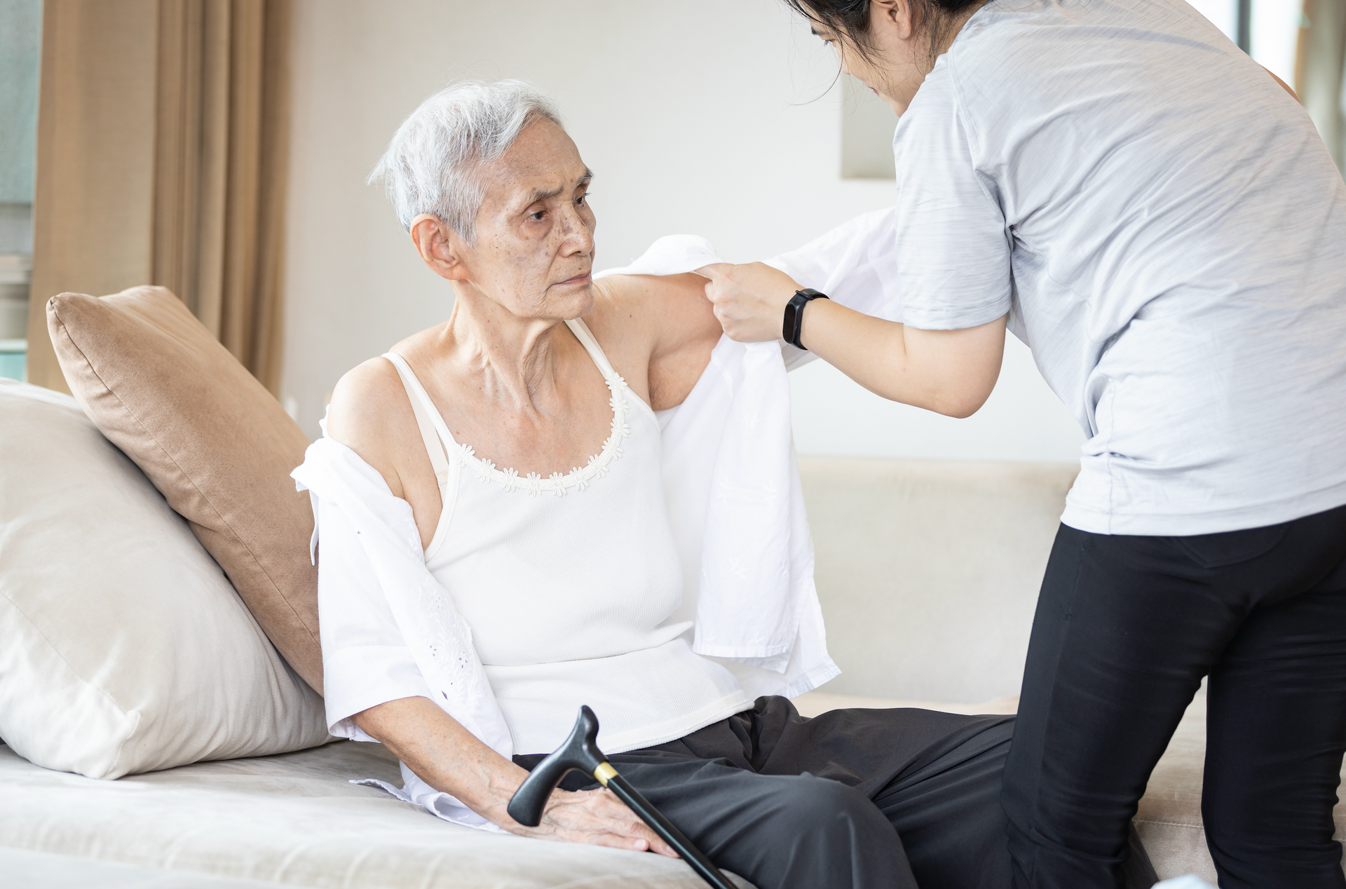 An elderly woman is sitting on a couch while a nurse helps her get dressed.