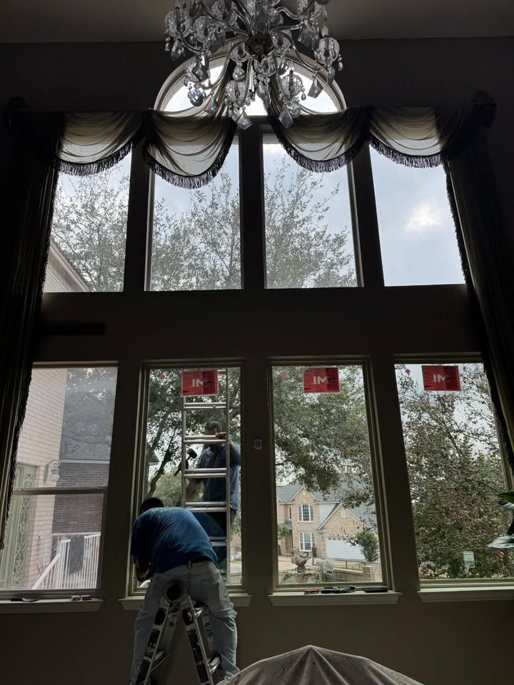 Person on a ladder working inside a room with tall windows and hanging drapes.