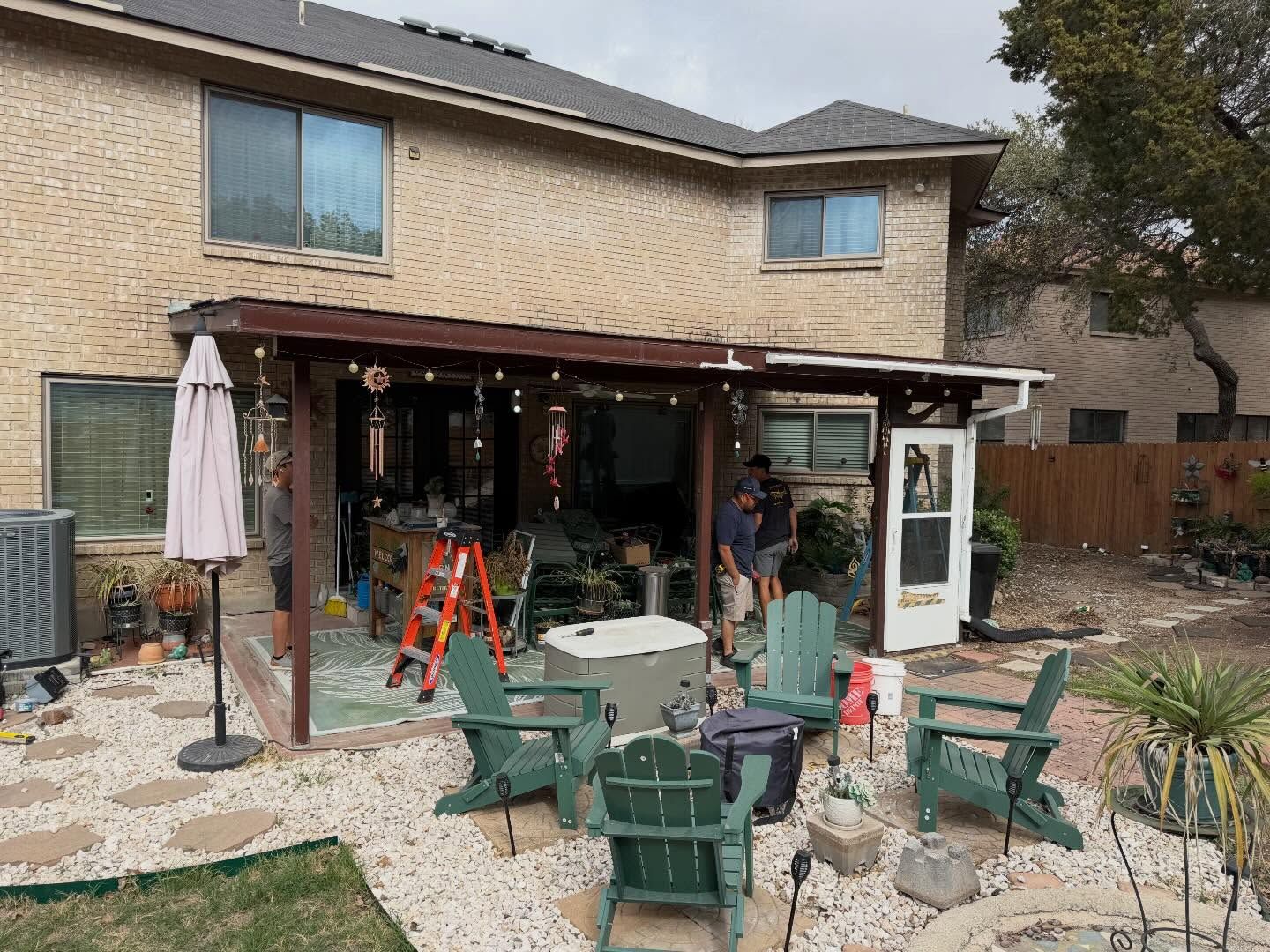 Backyard patio with green chairs, tables, umbrella, and a brick house with covered patio area