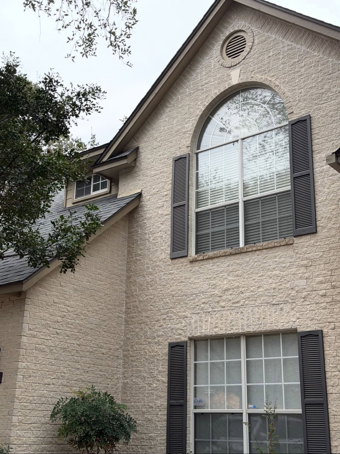 Beige stucco house exterior with arched window, dark shutters, and tree branches overhead