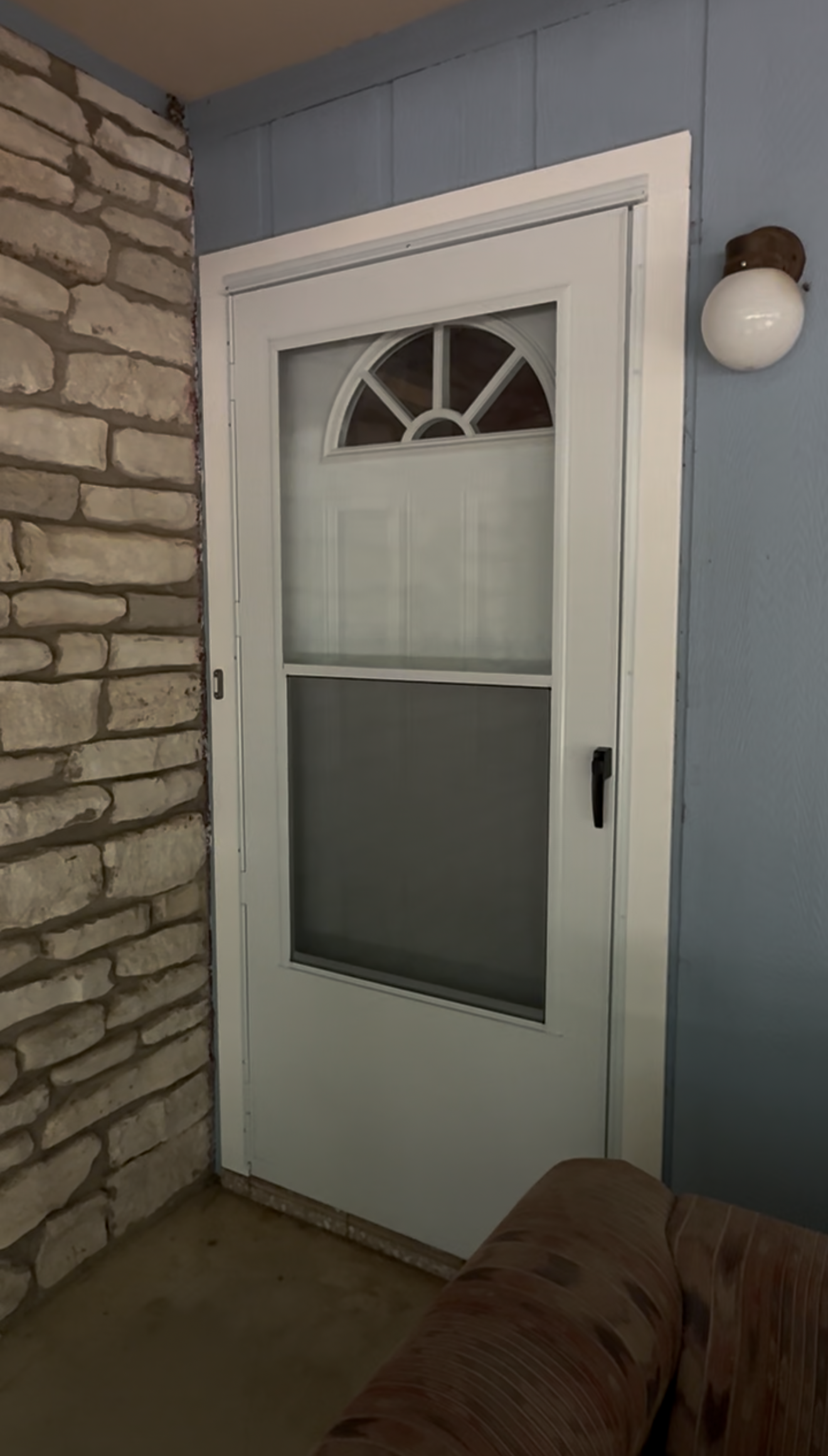 Front door with frosted glass, white trim, stone wall, and wall-mounted light on a porch.