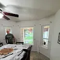 Bright unfinished room with white walls, exposed ceiling fan, and two windows showing a green yard.