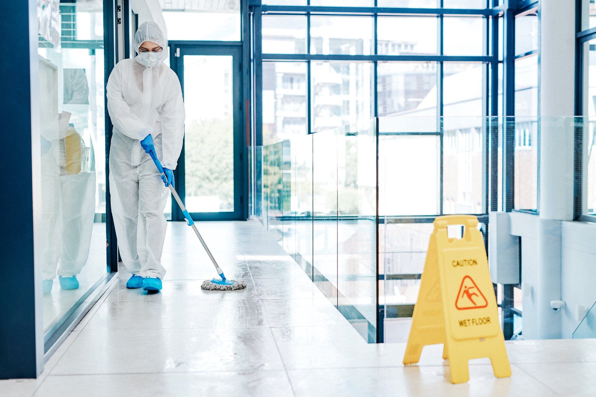 A man in a protective suit is mopping the floor of a building next to a caution sign.