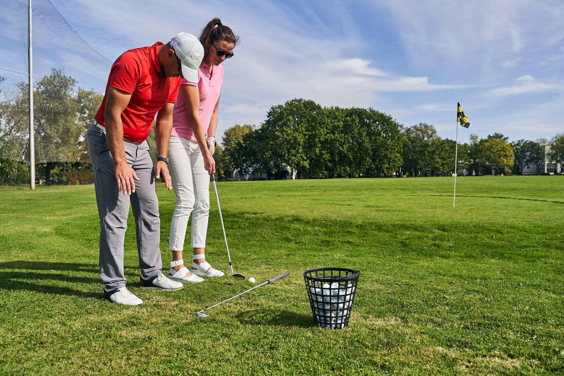 Man coaching woman on a golf course, reviewing swing. Sunny day, green grass.