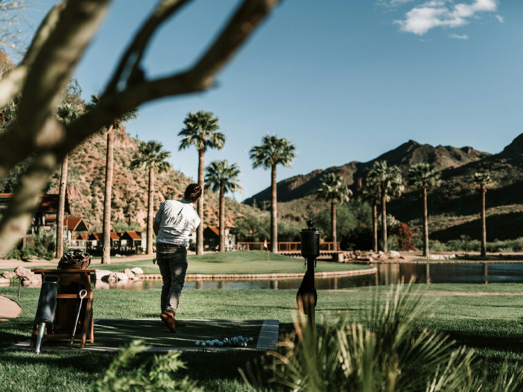 Golfer swinging on a green, palm trees, lake, and mountains in the background under a sunny sky.