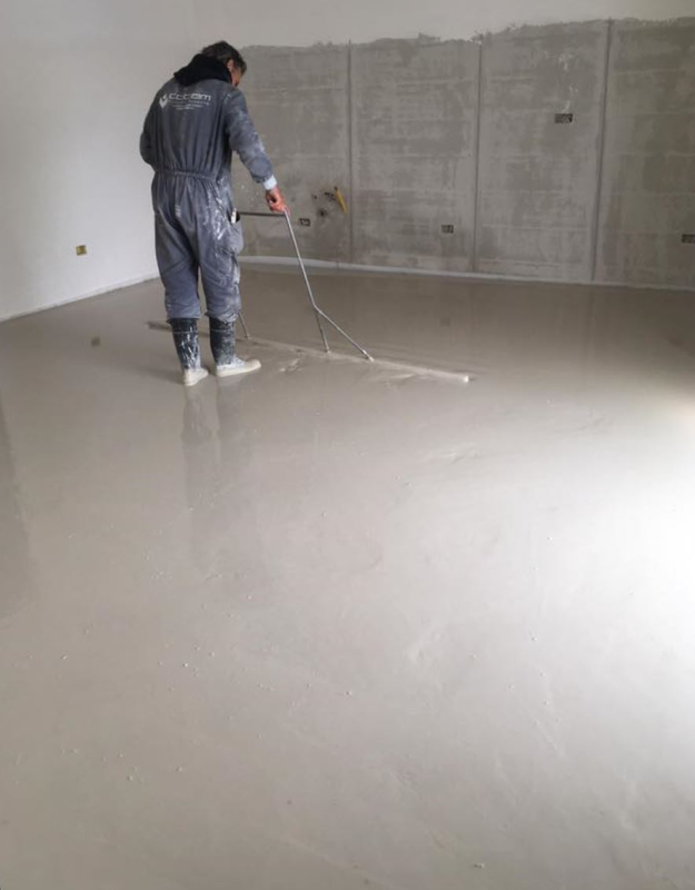 Una persona utilizza un attrezzo a manico lungo per levigare un pavimento di cemento grigio in una stanza spoglia.