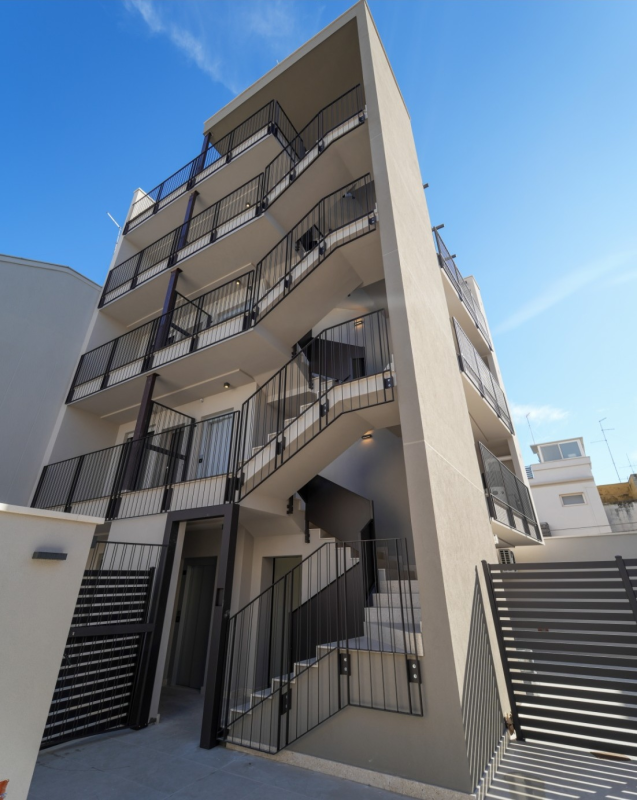 Esterno di un moderno condominio con scale esterne e ringhiere nere sotto un cielo azzurro.