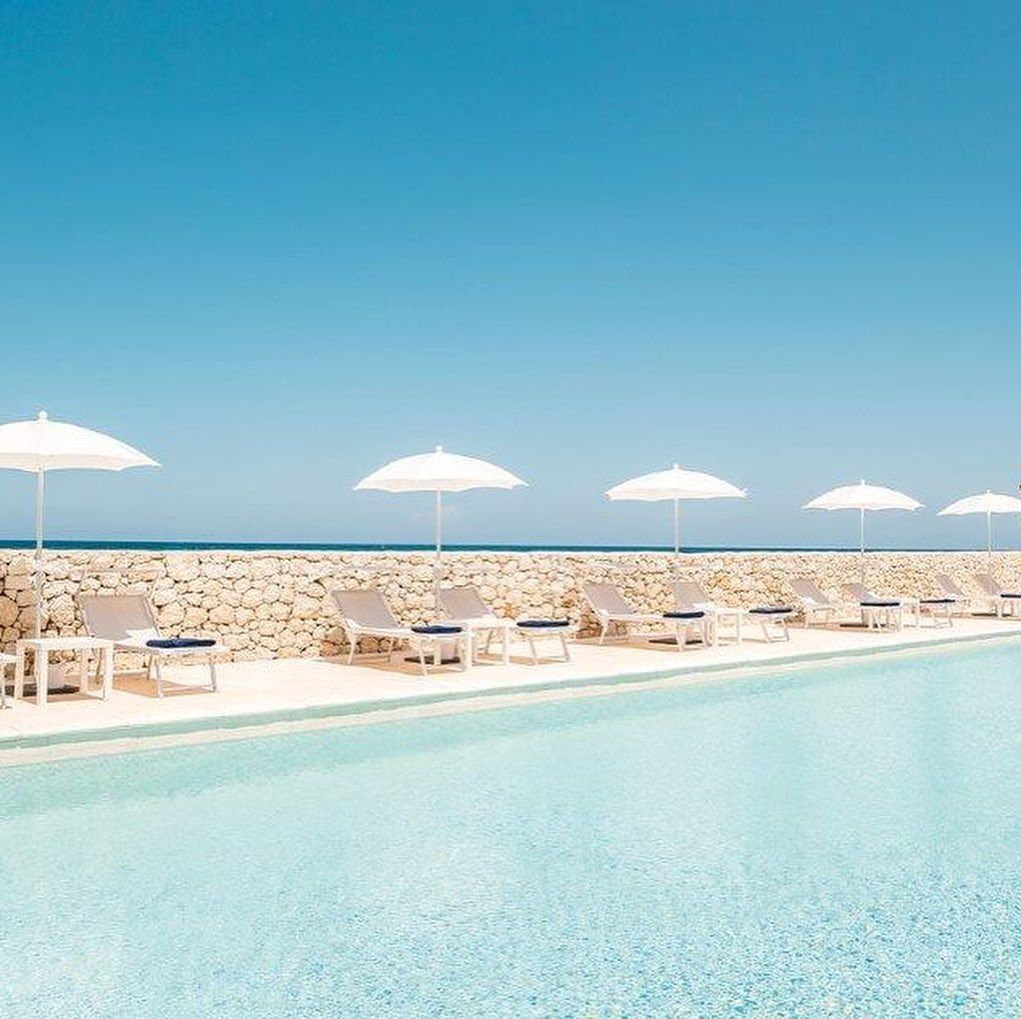 Piscina soleggiata in riva al mare con ombrelloni bianchi, lettini e un orizzonte blu sull'oceano.