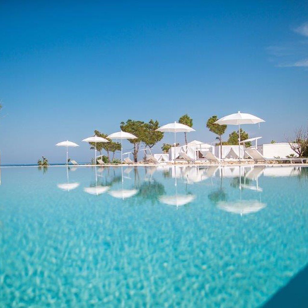 Piscina a sfioro turchese con ombrelloni bianchi e lettini sotto un cielo azzurro limpido.