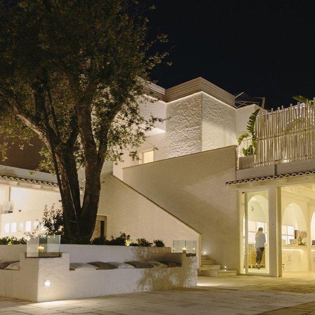 Cortile moderno illuminato di notte con edifici bianchi, un grande albero e ingressi ad arco.