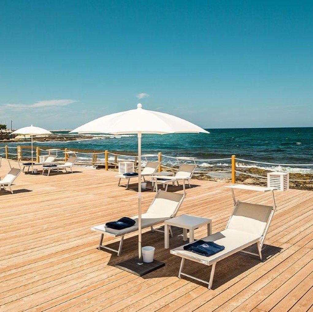 Sedie a sdraio e ombrellone bianco su una terrazza in legno in riva al mare, sotto un cielo azzurro e limpido.