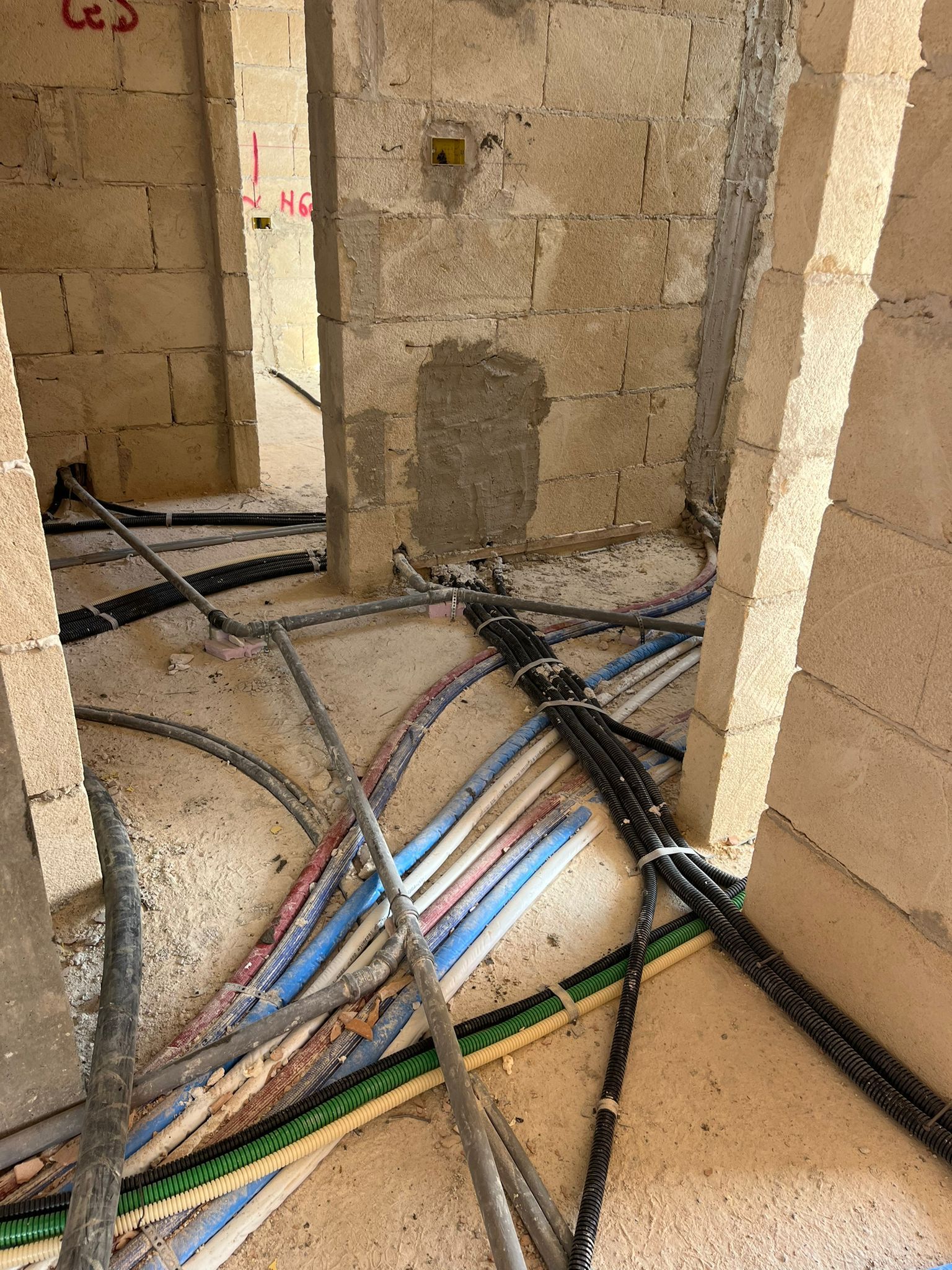 Cantiere edile con cavi elettrici a vista che corrono su un pavimento polveroso tra muri di blocchi non rifiniti.
