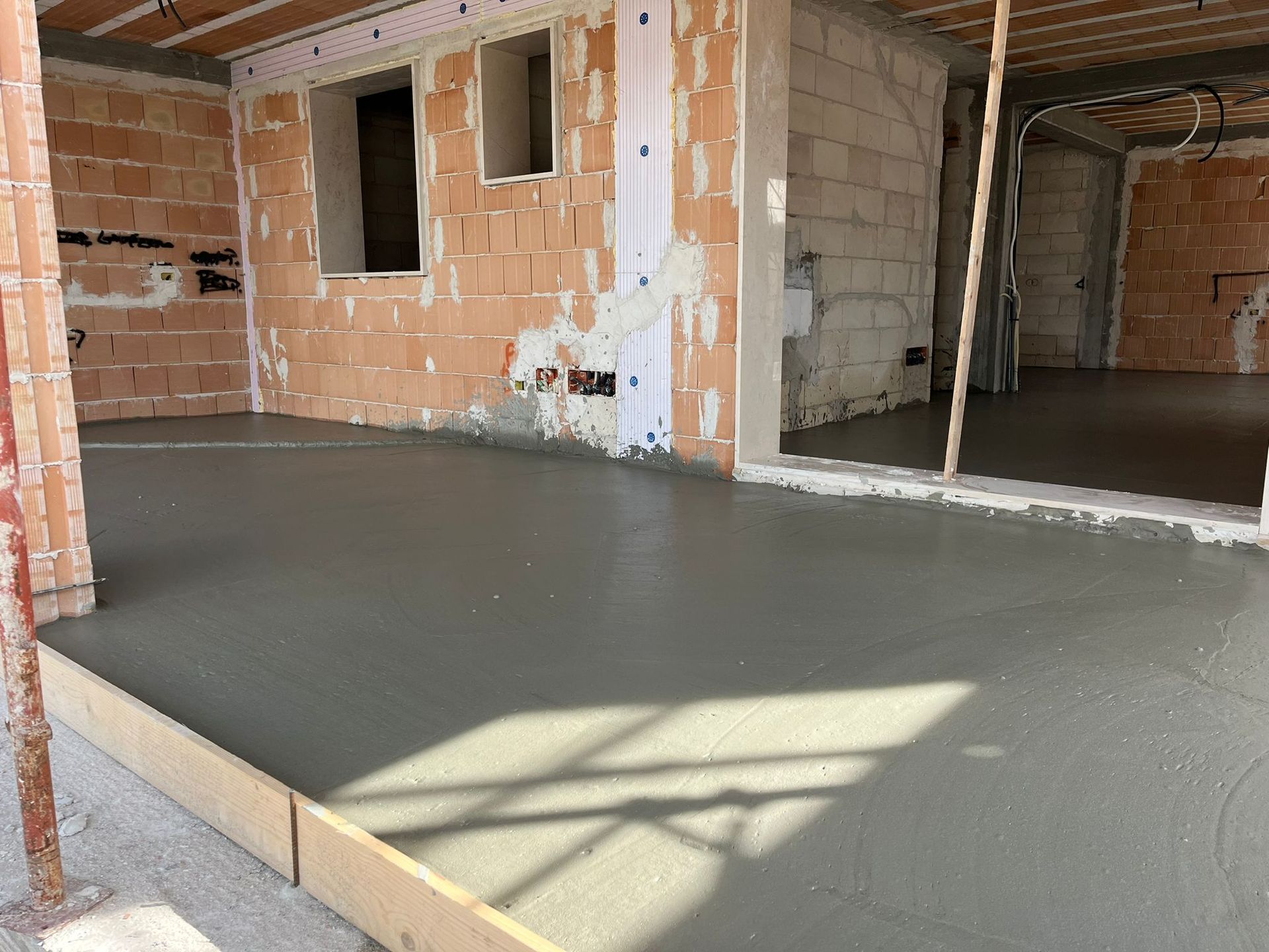 Pavimento in cemento grigio appena gettato all'interno di una stanza in mattoni non rifiniti con porte aperte.