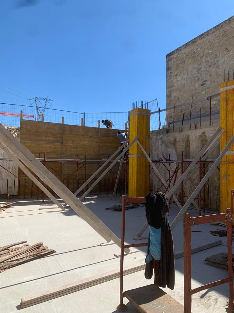 Cortile di un cantiere edile con impalcature, travi e una giacca nera appesa sotto un cielo azzurro limpido.