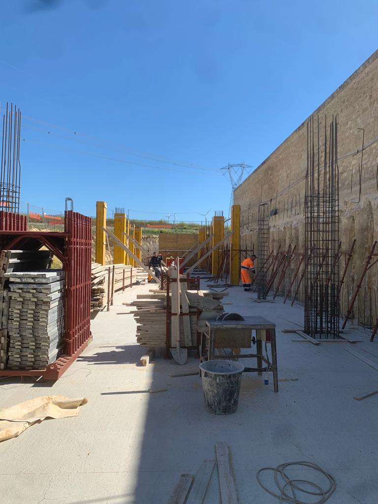 Corridoio di un cantiere edile con operai, impalcature, barre d'armatura e attrezzi sotto un cielo azzurro e limpido.