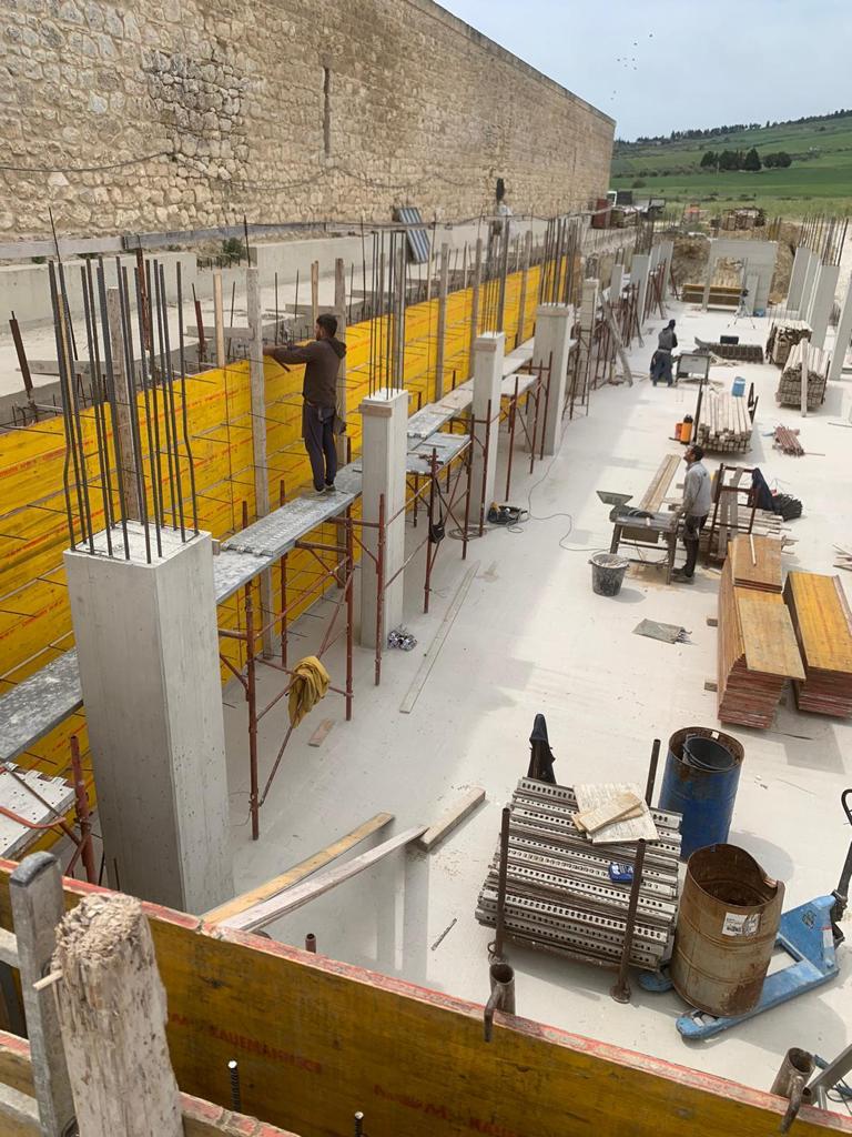 Cantiere edile con muri in cemento, casseforme gialle, barre d'armatura e operai intorno a ponteggi e materiali.