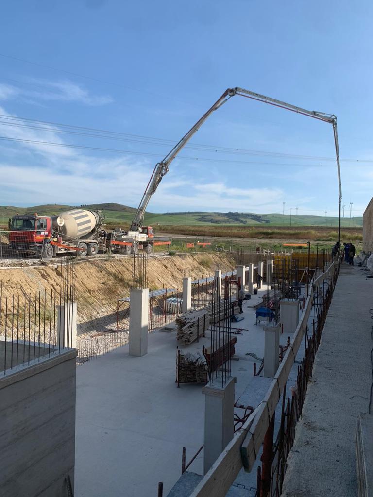 Cantiere edile con fondamenta in cemento, barre d'armatura e un'autobetoniera sotto un cielo azzurro.