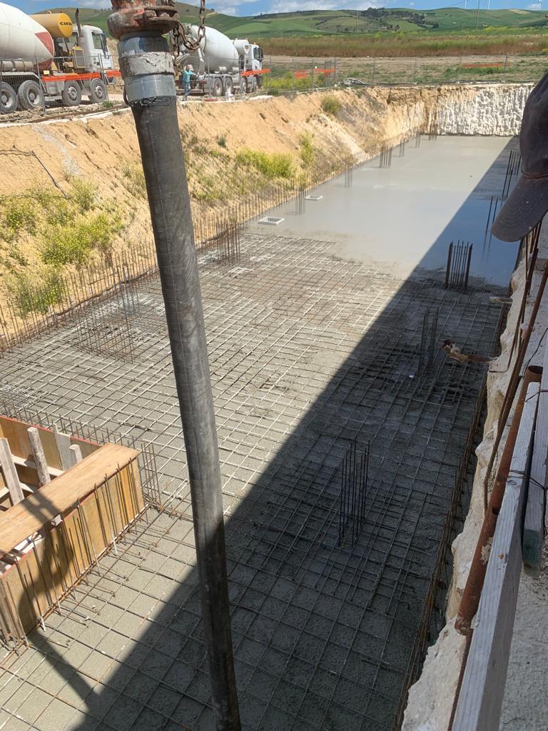 Cantiere edile con reti di barre d'acciaio accanto a un canale fangoso e braccio di escavatore in primo piano.