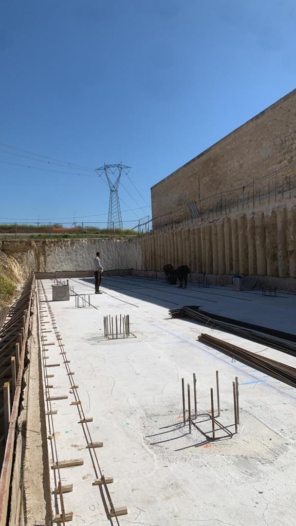 Cantiere per fondamenta in cemento armato, muro di contenimento e alti cavi elettrici sotto un cielo sereno.