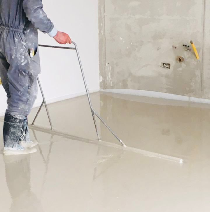 Un operaio liscia un pavimento di cemento fresco con un lungo attrezzo in una stanza non ancora rifinita.