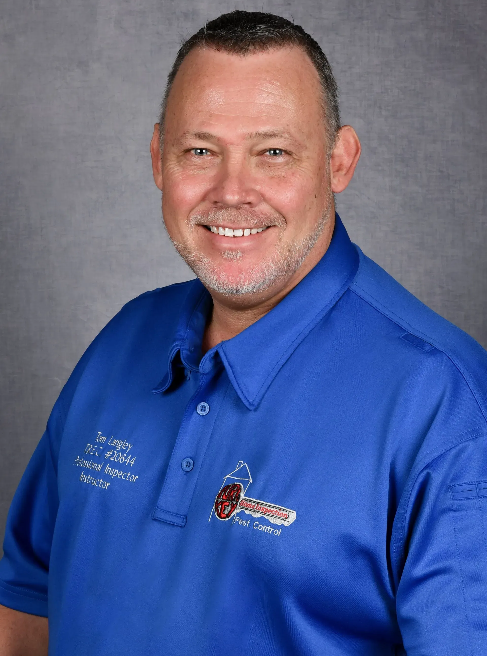A person smiling, wearing a blue collared shirt with an embroidered logo, set against a grey background.