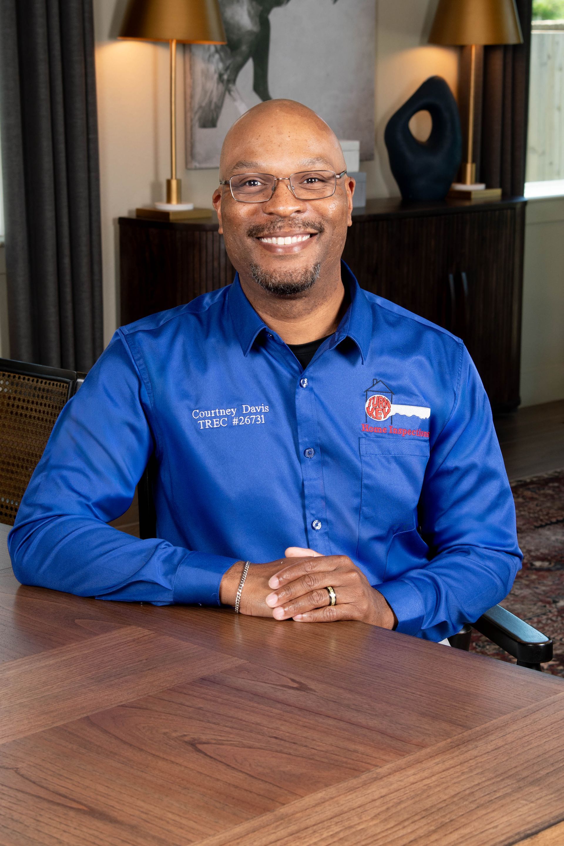 A person smiling, wearing a blue collared shirt with an embroidered logo, set against a grey background.