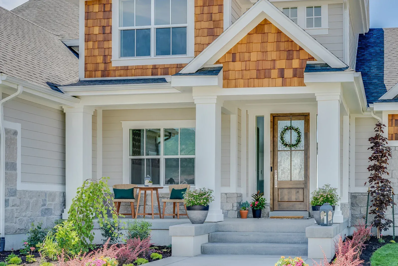 A modern home entrance with white columns, light-colored siding, wood shake accents, a wooden front door, and porch chairs.