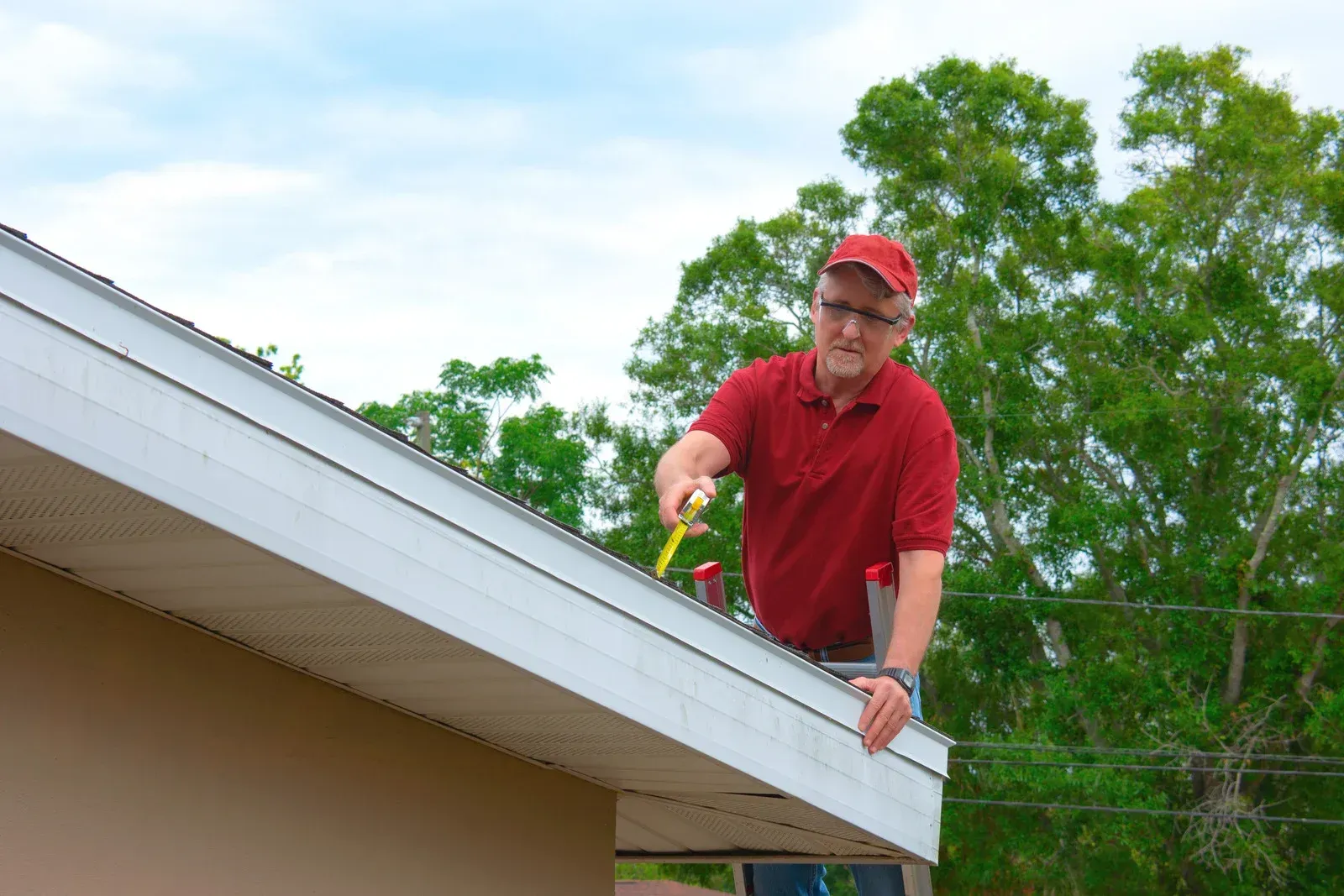 A person in a red polo shirt and cap using a tool to work on the edge of a house roof while standing on a ladder.