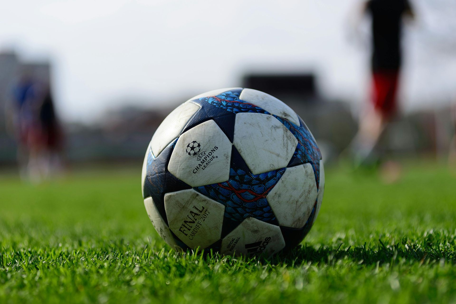 A Champions League soccer ball rests on a grassy field, with blurred figures visible in the background.