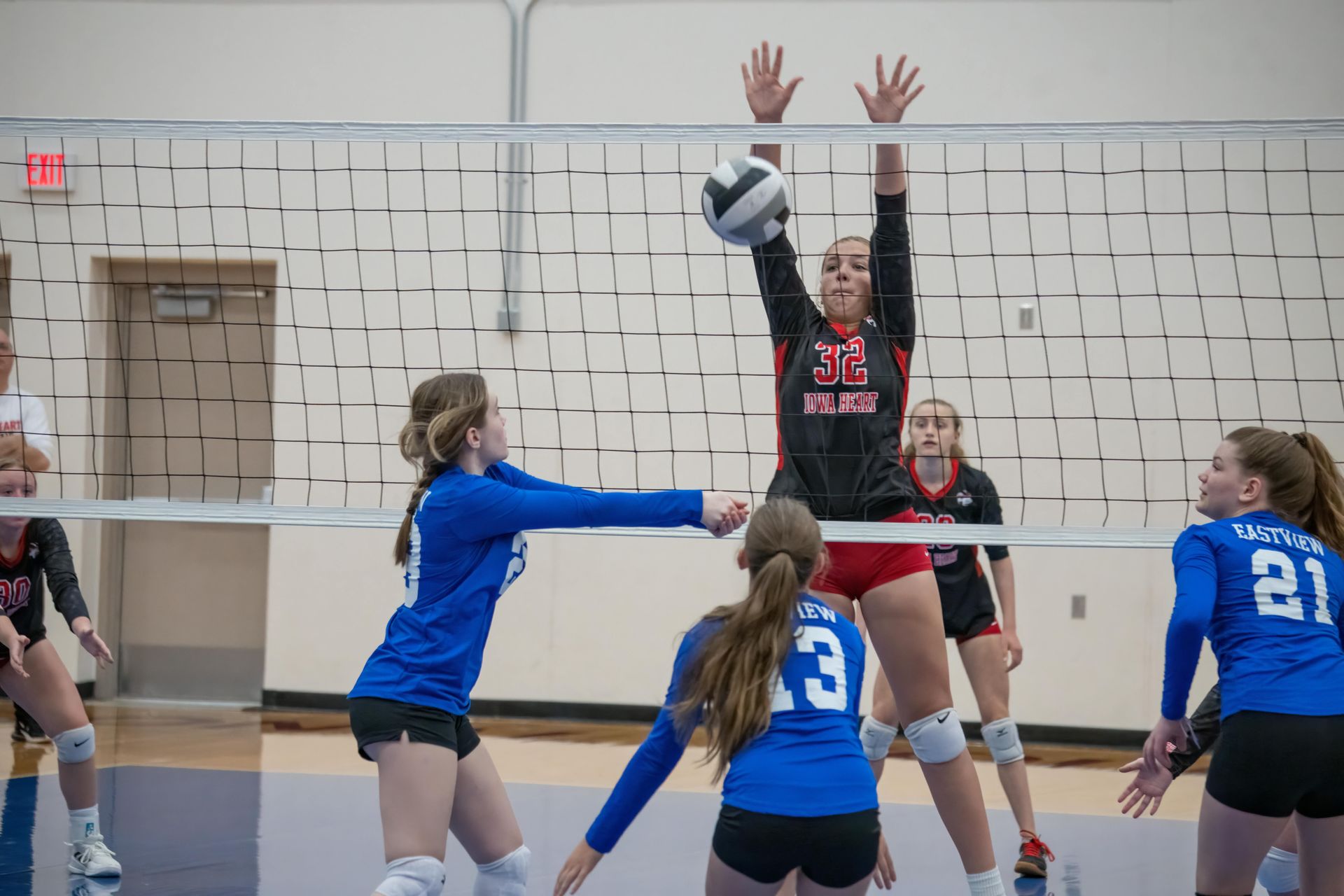 Volleyball players in blue and black uniforms compete at the net during an indoor match.