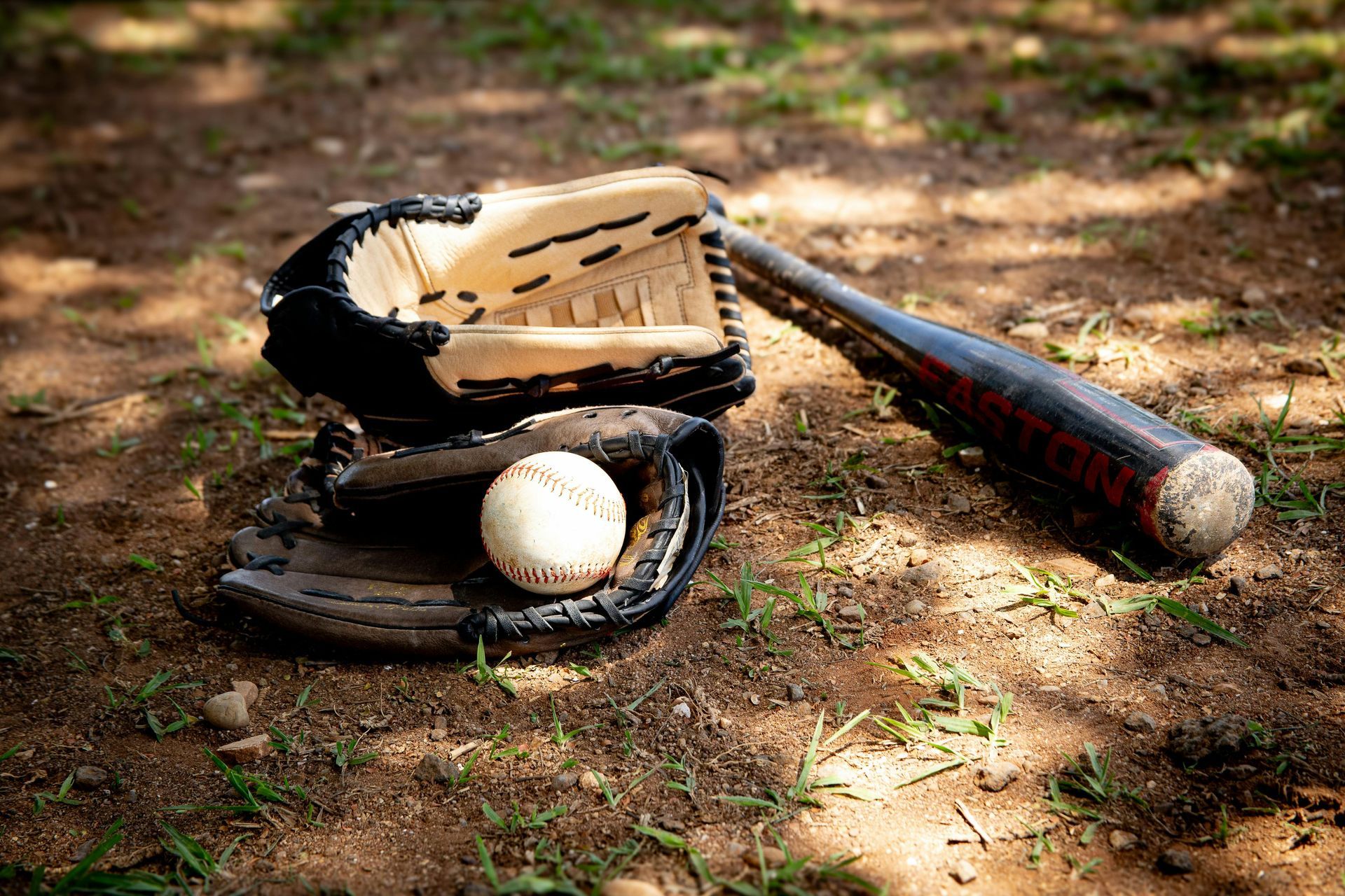 A baseball glove holding a baseball sits on a dirt field next to a dark baseball bat.