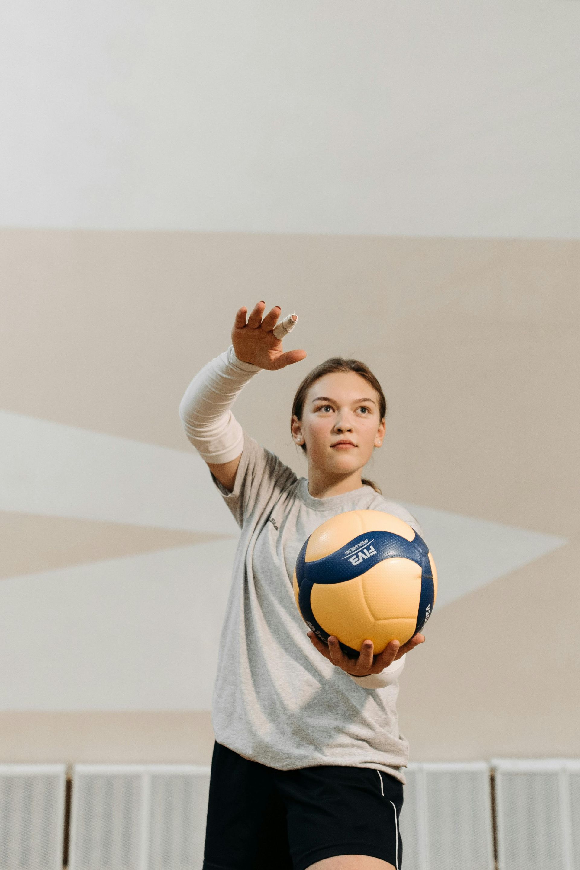 A person in a gray shirt and black shorts holds a yellow and blue volleyball while preparing to serve in an indoor gym.